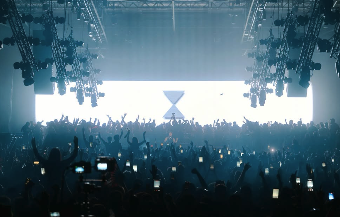 A packed electronic music dancefloor beneath stage lights